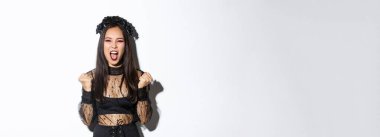 Neşeli, çekici Asyalı cadı imajı, Cadılar Bayramı kostümlü kadın sevinç ve yumruk pompasıyla bağırıyor, zaferi kutluyor, beyaz arka planda zafer kazanıyor..