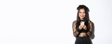 Gotik dantel elbiseli zarif bir esmer cadının resmi. Cep telefonu kullanıyor, kameraya gülümsüyor, beyaz arka planda duruyor..