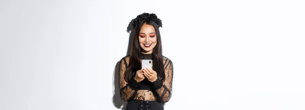 ハロウィンパーティーの魔女を装ったゴシックレースドレスの美しいアジアの女性のイメージ スマートフォンで立って白い背景に笑みを浮かべて — ストック写真