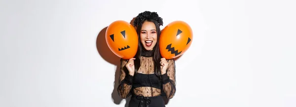 アジアの女の子のイメージで悪魔女の衣装を保持二つのオレンジ色の風船で怖い顔 ハロウィンを祝います 白い背景の上に立って — ストック写真
