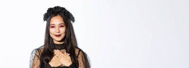 Minnettar gülümseyen Asyalı kadının yakın çekimi. Elleri göğsünde minnettar görünüyor. Beyaz arka planda cadı kostümü giyiyor..