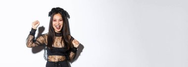 Cadılar bayramı partisinde eğlenen genç Asyalı bir kadının resmi. Neşeli ya da muzaffer olarak elini kaldırıyor. Cadı gotik elbisesi giyiyor, beyaz arka planda duruyor..