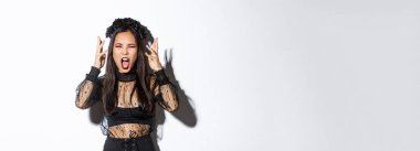 Image of angry asian woman in witch costume yelling at someone, looking mad, losing temper and screaming, standing over white background pissed-off.