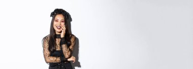 Gotik elbiseli ve siyah çelenkli mutlu Asyalı kadın imajı, Cadılar Bayramı partisinde gülüyor, beyaz arka planda duruyor..