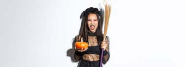 Cadılar bayramı partisi için cadı gibi giyinmiş, elinde süpürge ve balkabağı olan, beyaz arka planda duran Asyalı güzel bir kadın resmi..