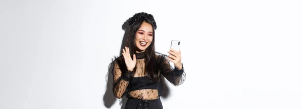 美しいとスタイリッシュなアジアの女性の肖像ゴシックレースのドレスと言っハイ ビデオ通話中にスマートフォンのカメラで手を振って 白い背景の上に立って — ストック写真