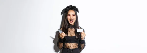 幸せな笑いアジアの女の子でハロウィンの衣装を身に着けていますアップビート 保持スマートフォンやクレジットカード 白い背景に立って — ストック写真