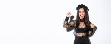 Cadılar bayramı partisinde eğlenen genç Asyalı bir kadının resmi. Neşeli ya da muzaffer olarak elini kaldırıyor. Cadı gotik elbisesi giyiyor, beyaz arka planda duruyor..