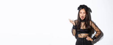 Frustrated and angry young asian woman in witch dress arguing with someone, looking pissed-off and bothered, shaking hand and looking scary at camera, standing over white background.
