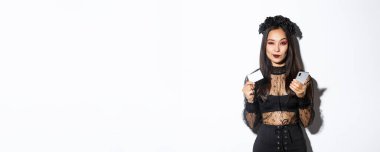 Şımarık genç kadın düşünceli görünüyor, kredi kartı ve cep telefonu tutuyor, internetten alışveriş yapıyor, beyaz arka planda duruyor..