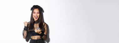 Mutlu Asyalı bir kadının güzel haberleri kutlaması, sevinçle yumruk tokuşturması, akıllı telefon tutması, cadılar bayramı kostümü giymesi, beyaz arka planda durması..