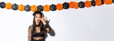 Cadı kostümü giymiş komik Asyalı kadın resmi, gotik dantel elbise giyiyor ve balkabağı şeritlerine karşı duruyor, cadılar bayramı süslemeleri yapıyor..