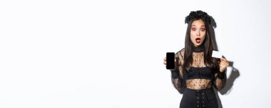 Cadı kostümlü Asyalı kız kredi kartını cep telefonu ekranında gösteriyor ağzı açık, şaşırmış, cadılar bayramını kutluyor, beyaz arka plan.