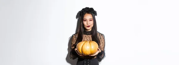 黒のドレスで狡猾なアジアの女性のイメージ ハロウィンに邪悪な魔女を装った 大きなカボチャを保持します 白い背景の上に立って — ストック写真