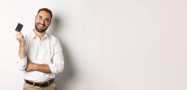 Alışveriş yapmayı düşünen ve kredi kartı tutan yakışıklı bir adamın görüntüsü. Sol üst köşeye bakıyor. Düşünceli, beyaz arka plan..