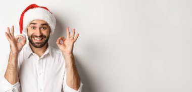 Parti, kış tatili ve kutlama konsepti. Neşeli adam Noel 'den zevk alıyor ve iyi bir işaret veriyor, gülümsüyor, Noel Baba şapkası takıyor, beyaz arka plan.