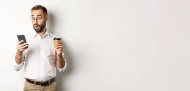 Kahve içen ve cep telefonundaki mesaja şaşıran bir iş adamı beyaz arka planda şaşkın şaşkın dikiliyor..