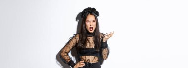 Frustrated and angry young asian woman in witch dress arguing with someone, looking pissed-off and bothered, shaking hand and looking scary at camera, standing over white background.