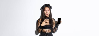 Siyah gotik elbiseli sürpriz Asyalı kız, nefes nefese eğleniyor ve cep telefonu ekranını işaret ediyor, cadılar bayrağı ya da tanıtımını gösteriyor, beyaz arka planda duruyor..