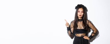 Cadı kostümlü, çekici Asyalı kadın, sol üst köşesini işaret ediyor, cadılar bayramı promosyonuyla ilgileniyor, beyaz arka planda duruyor ve düşünüyor.