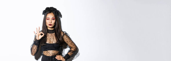 Sassy beautiful asian woman in witch costume showing okay gesture, all under control. Confident girl in halloween gothic dress like or approve something, standing white background.