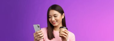 Yakın çekimde ihale dostu Asyalı kadın blogcu sosyal medyayı kontrol ediyor hazır kahve içiyor, neşeli bir şekilde gülümsüyor telefon ekranına bakıyor, ilginç makaleler okuyor, mesajlaşıyor, mor arka plan okuyor..
