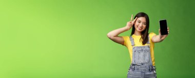 Akıllı telefon ekranında başvuru gösteren kaygısız Asyalı esmer, zafer barış işareti yapıyor, neşeyle gülümsüyor, sosyal medya popülaritesiyle övünüyor, takipçi sayısı, yeşil arka planda duruyor.