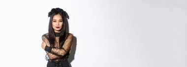 Şık gotik elbise ve çelenk giymiş genç zarif bir kadın, vücudunu kucaklıyor ve kameraya küstah bakıyor, cadılar bayramı kostümü giymiş, beyaz arka planda duruyor..