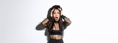 Gotik dantelli elbise ve çelenk giymiş, pusuya düşmüş, cadılar bayramı kostümü giymiş, nefes nefese kalmış ve endişeli görünen, beyaz arka planda duran Asyalı kadın görüntüsü..