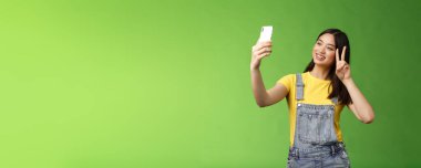Tatlı Asyalı esmer kadın, akıllı telefonu tut, poz ver, telefon ekranına bak, fotoğraf çek, zafer barış işareti göster, yurtdışında neşeyle sohbet et, yeşil arka planda dur. Boşluğu kopyala