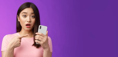 Endişeli, kafası karışık, genç Asyalı bir kadın akıllı telefonu açık tutuyor, parmağını telefona doğrultuyor, sorgulanıyor gibi görünüyor, üzgün görünüyor, garip yazılar görüyor, mor arka planda duruyor. Boşluğu kopyala
