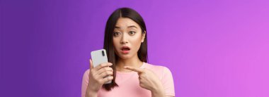 Endişeli, kafası karışık, genç Asyalı bir kadın akıllı telefonu açık tutuyor, parmağını telefona doğrultuyor, sorgulanıyor gibi görünüyor, üzgün görünüyor, garip yazılar görüyor, mor arka planda duruyor. Boşluğu kopyala