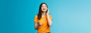 Kaygısız, sevimli Asyalı kız harika müziğin keyfini çıkar favori şarkıları dinle, kulaklıklara dokun zevkten gözlere dokun, eşlik et, akıllı telefonları tut mavi arka plan.