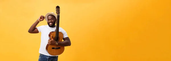 Muscular black man playing guitar, wearing jeans and white tank-top. Isolate over yellow background