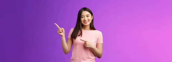 陽気な優しいアジアの笑顔の女の子が右を指して プロモーションを示し 新しい製品を紹介し 喜んで現在のクールな広告を成長させ ピンクのTシャツ紫の背景が面白がって発信立って — ストック写真
