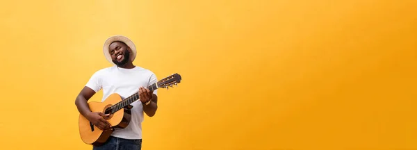 Muscular black man playing guitar, wearing jeans and white tank-top. Isolate over yellow background