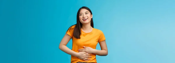 Happy staffed cute asian girl feelings satisfaction after eating favorite food, smiling broadly rubbing tummy, touch stomach from delight and joy, like eat delicious meals, stand blue background.