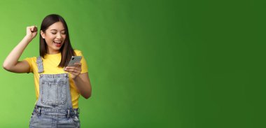 Güzel, neşeli Asyalı kız, akıllı telefonu tut, iyi haberleri kutla, iyi haberleri oku, mutlu bir şekilde gülümseyen telefon ekranına bak, başarıya ulaş, izin al.