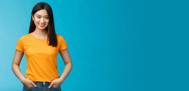 Kendinden emin gülümseyen Asyalı kadın mavi arka planda duruyor kamera, kot pantolonun cepleri kendine güvenen titreşimler, olumlu sonuçların tadını çıkar cilt bakımı prosedürleri, sarı tişört giy.