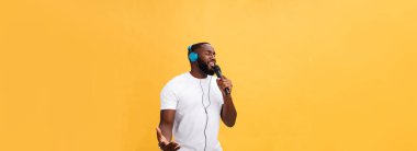 Neşeli pozitif şıklığın portresi. Yakışıklı Afrikalı bir adam mikrofon tutuyor ve kulaklıkları başında müzik dinliyor sarı arka planda haftasonu tatilinin tadını çıkarıyor..