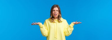 What problem wtf. Pissed-off frustrated and angry young blond caucasian girl shrugging with confused annoyed expression, cant understand what happened, dont know anything, blue background.