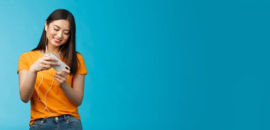 Hoş görünümlü şehir kızı tatilini yarış oyunu oynayarak geçiriyor, akıllı telefonu yatay pozisyonda tutuyor telefonu hareket ettiriyor, geniş bir coşkuyla gülümsüyor mavi arka plan..
