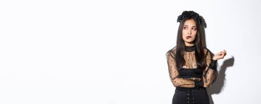 Cadı kostümlü düşünceli güzel Asyalı kadın dudak ısırıyor ve sol üst köşeye bakıyor. Cadılar bayramı elbisesi içinde beyaz arka planda dururken hayal kuruyor..