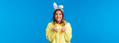 Tatiller, gelenekler ve kutlama konsepti. Sevimli ve aptal gülümseyen sarışın kadın kameraya bakıyor ve kafasında sahte kulaklar olan tavşanı taklit ediyor, mavi arka planda duruyor..
