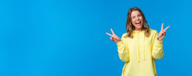 Hadi biraz eğlenelim. Kaygısız, neşeli, dost canlısı kawaii görünümlü sarı kapüşonlu sarışın kız, göz kırpan ve gülümseyen beyaz dişler sevimli barış jestleri, mavi arka plan.