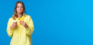 Akıllı telefon ekranından fotoğrafına bakan kız, iyi bir yüz ifadesiyle, memnun ve tasvip edilmemiş görünüyor. Mobil uygulamalarda fotoğraf düzenledikten sonra, kağıt bardaktan kahve içtikten sonra..