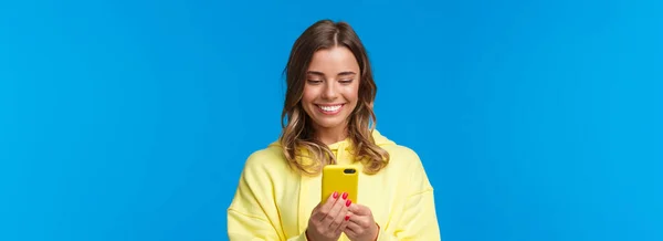 特写镜头 傻傻的金发欧洲女人 穿着黄色的帽衫 在手机展示会上笑着看可爱的视频 和男朋友聊天 站在蓝色的背景上 — 图库照片