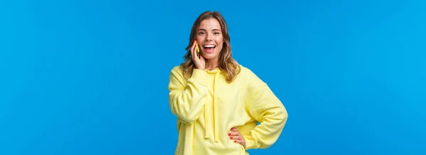 見た目の良いヨーロッパのブロンドの女の子の黄色のパーカーは 携帯電話で活発な話 バーで友人のための予約テーブルとして笑顔カメラ 注文食品配達 青の背景 — ストック写真
