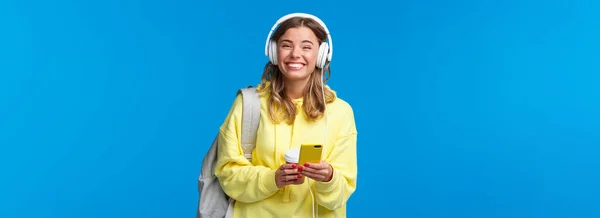 快乐的年轻女生 背着背包 听着耳机里最喜欢的演奏者 无忧无虑地笑着看着相机 拿着手机 站在蓝色的背景上 — 图库照片