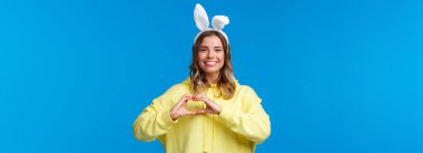 Tatiller, gelenekler ve kutlama konsepti. Tavşan kulaklı karizmatik sarışın kızın belden yukarı portresi, Paskalya tatili ile herkesi tebrik et, kalp hareketi ve gülümseme göster.